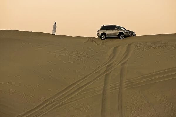 Man in traditional Arab dress looking at 4x4 car stuck on the ridge of a dune during a desert safari near Abu Dhabi, United Arab Emirates