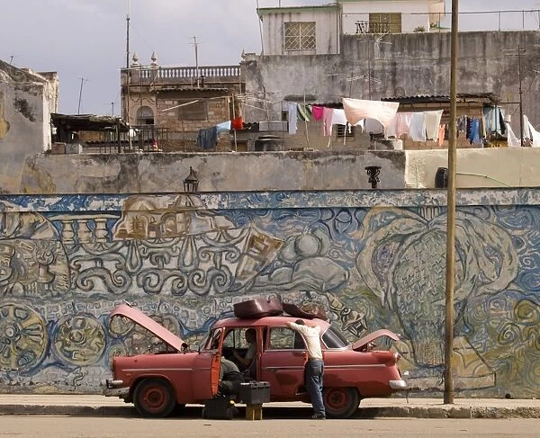 Two men repairing an old car parked in front of a colourful wall mural in central Havana