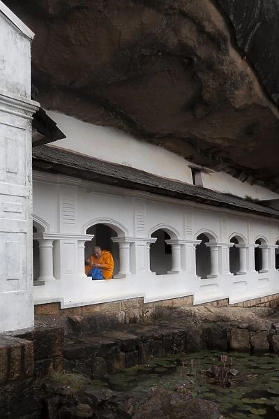 A monk sits and looks at his phone beneath shelter, Royal Rock Temple, Golden Temple of Dambulla, UNESCO World Heritage Site, Dambulla, Sri Lanka, Asia