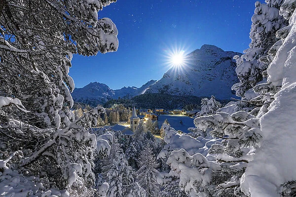Moon glowing over the snowy bell tower of Chiesa Bianca and woods in winter, Maloja, Bregaglia, Engadine, Canton of Graubunden, Switzerland, Europe