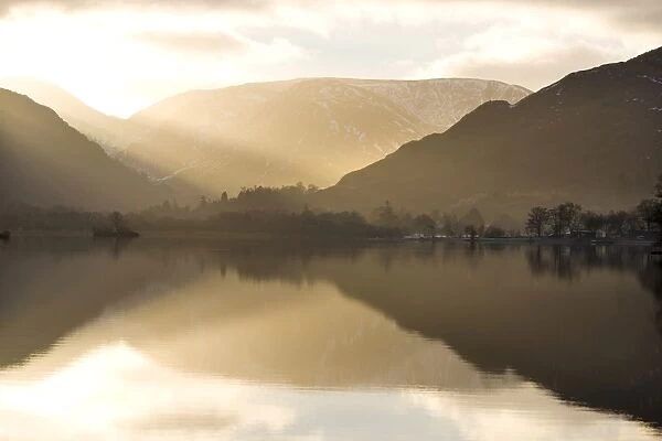 Morning sunlight bursting through clouds over fells with reflections in Lake Ullswater