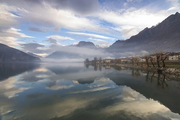 Mountains and village are reflected in Lake Mezzola at dawn shrouded by mist, Verceia