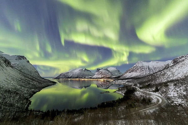 Northern lights (aurora borealis) and stars light up the snowy peaks reflected in the cold sea