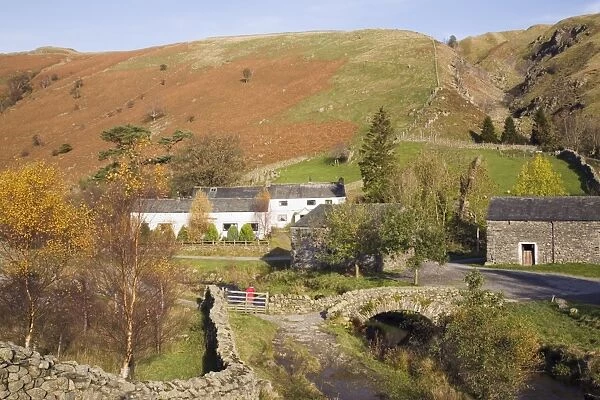 Old stone packhorse bridge over Watendlath Beck with dry stone wall and farm buildings in picturesque village in autumn, Watendlath, Lake District National Park, Cumbria, England, United