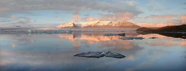 Panoramic view across the calm water of Jokulsarlon glacial lagoon towards snow-capped mountains and icebergs bathed in late afternoon light in winter, at the head of the Breidamerkurjokull Glacier on the edge of the Vatnajokull National Park, South Iceland, Iceland, Polar Regions