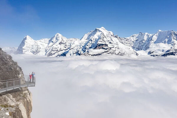 Two people photographing snowcapped Eiger and Monch peaks in fog from elevated walkway
