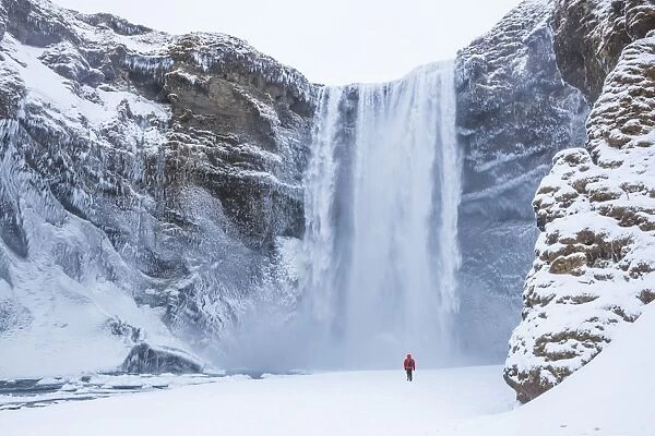 One person in red jacket walking in the snow towards Skogafoss waterfall in winter
