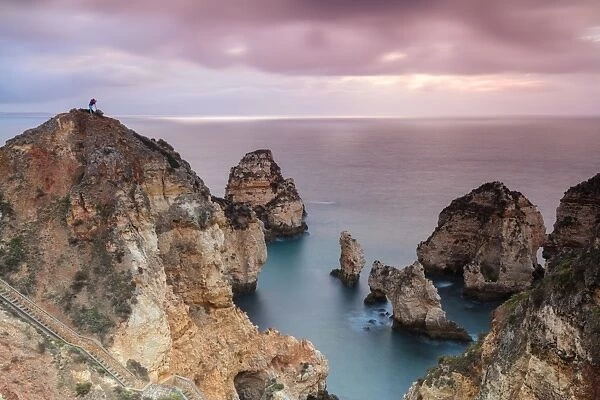 Photographer on top of cliffs surrounded by sea under the pink sky at sunrise, Ponta Da Piedade