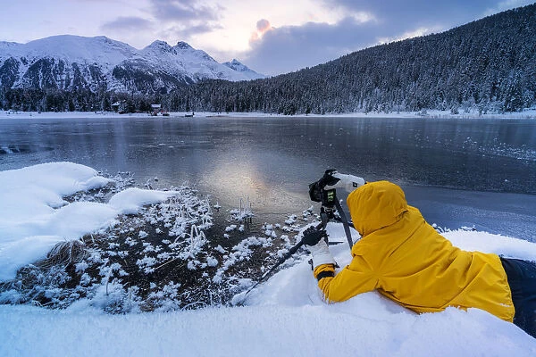 Photographer taking pictures of frozen Lej da Staz at dawn lying on the snow, Engadine