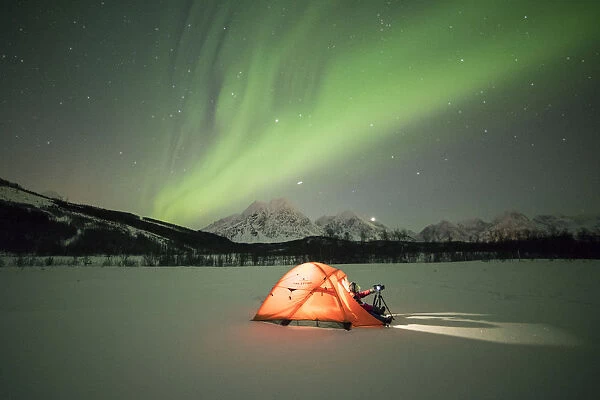 Photographer in a tent lit up by Northern Lights (aurora borealis) and starry sky