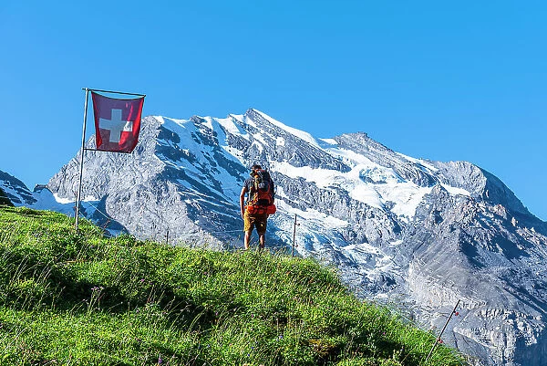 Rear view of a man with backpack hiking the Swiss Alps passing a Swiss flag flying, Oeschinensee, Kandersteg, Bern Canton, Switzerland, Europe