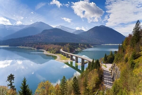 Reflections of a road bridge over Lake Sylvenstein, with mountains in the background