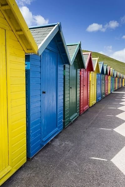 Row of colourful beach huts and their shadows with green hill backdrop, West Cliff Beach