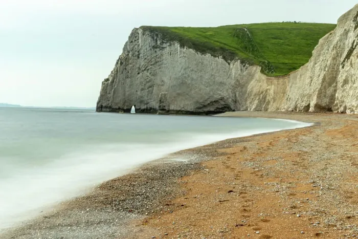 Framed Print : Serene beach with smooth waters, pebbles, and a towering cliff under a cloudy sky at Durdle Door, Jurassic Coast, UNESCO World Heritage Site, Dorset, England, United Kingdom, Europe