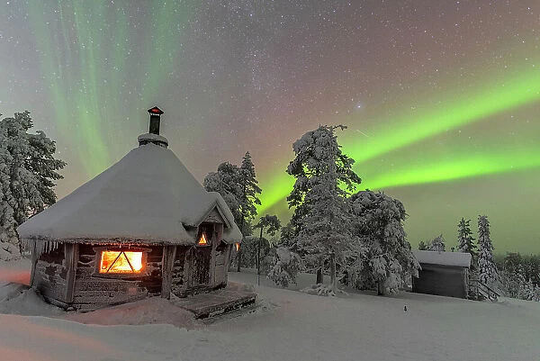 Shades of green color the Northern Lights (Aurora Borealis) above a snowy landscape with a typical hut lit by a fire inside in the foreground, Finnish Lapland, Finland, Europe