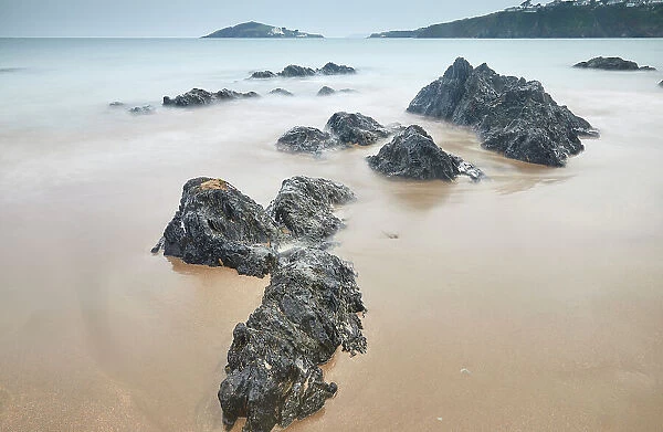 Shoreline boulders buried in sand face an advancing tide at dusk, looking from Bantham beach towards Burgh Island, on the south coast of Devon, England, United Kingdom, Europe