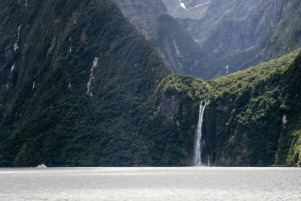 A sightseeing ship dwarfed by a tall waterfall in a fjord, South Island, New Zealand