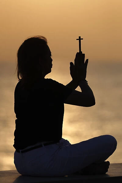 Silhouette of faithful woman praying with Christian cross at sunset as concept for