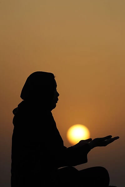 Silhouette of a Muslim woman in abaya praying with her hands at sunset