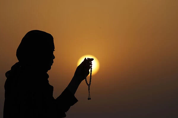 Silhouette of a Muslim woman holding prayer beads in her hands and praying at sunset