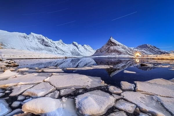 Star trails and lights on the snowy peaks reflected in the cold sea, Volanstinden