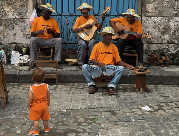 Street band wearing orange shirts playing music on the pavement watched by toddler wearing orange clothes, Habana Vieja (Old Havana), Havana, Cuba, West Indies