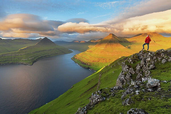 Summer view of a hiker standing on top of a mountain overlooking the fjord at sunrise, Funningur fjord, Eysturoy island, Faroe islands, Denmark, Europe