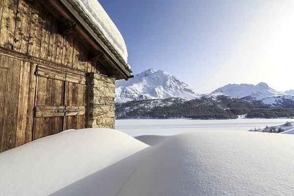 The sun, covered by thin clouds, illuminating a typical hut covered with snow at the Maloja Pass, Graubunden, Swiss Alps, Switzerland, Europe