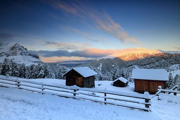 The sunrise lights up the snowy woods and huts, Passo Delle Erbe, Funes Valley, South Tyrol