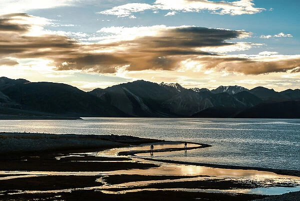 Sunset at Pan Gong lake with golden light reflection in the flat waters showing two people walking along the shore, Ladakh, northern India, Asia
