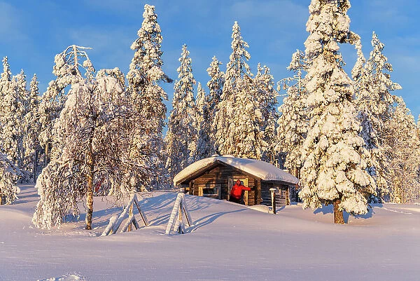 Tourist in the early morning sun stands in front of an isolated chalet in the snowy forest, Norrbotten, Swedish Lapland, Sweden, Scandinavia, Europe