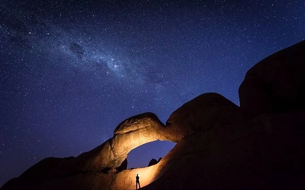 Tourist under the Rock Arch bridge in the Spitzkoppe region with a view of the Milky Way
