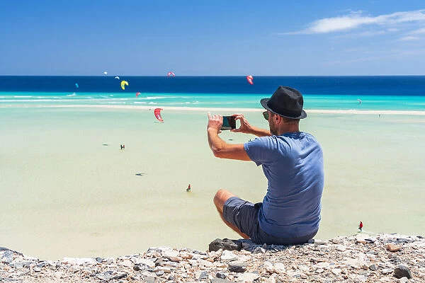Tourist with straw hat photographing kiteboarders with smartphone at Sotavento beach