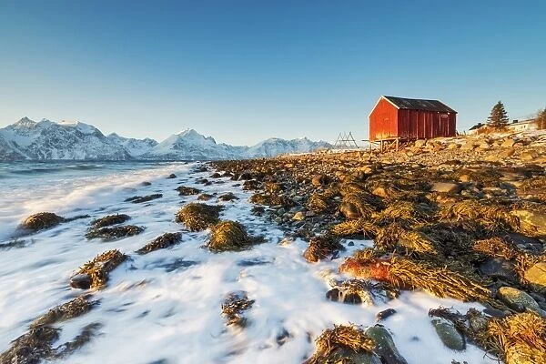 Typical wooden hut called Rorbu surrounded by waves of the cold sea and snowy peaks