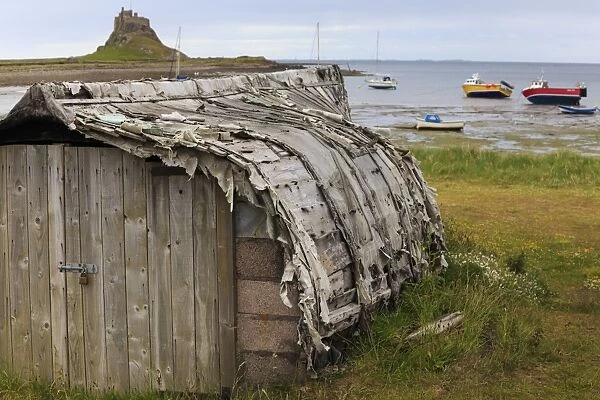 Upturned weathered boat hut with Lindisfarne Castle and fishing boats at low tide