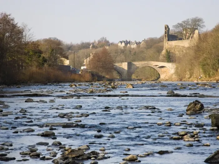 Framed Print : View along the boulder-strewn River Tees to the ruined castle and 16th century County Bridge