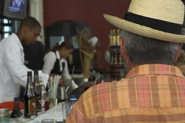 Back view of a man in a straw hat sitting at a bar, with bar staff in the background