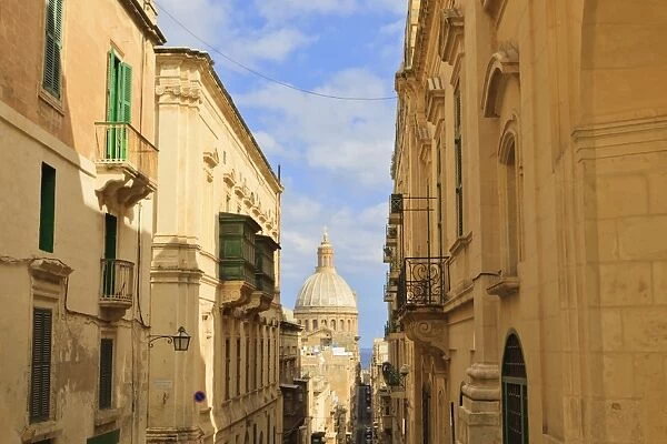 View down a narrow street with golden stone, green shutters and balconies, to the Carmelite Church and its dome, Valletta, Malta, Europe