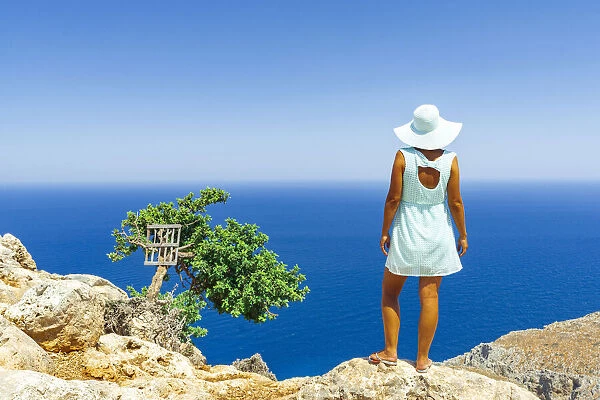 Back view of woman with fashion dress and hat looking at the sea from cliffs, Crete