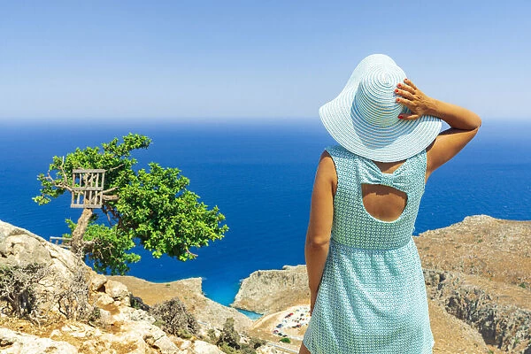 Back view of young woman with dress and sun hat watching the sea, Crete, Greek Islands