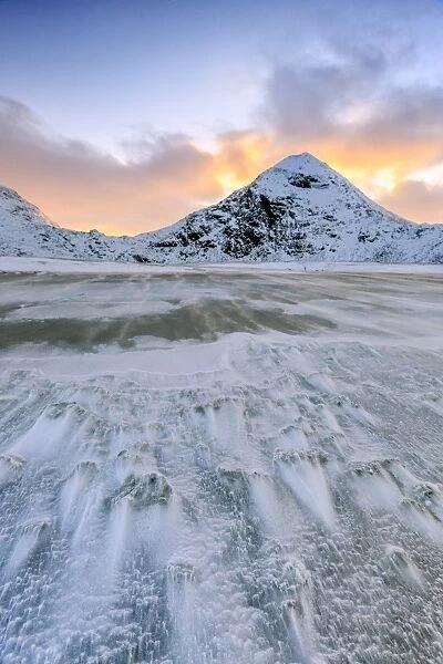 Wave advances towards the shore of the beach surrounded by snowy peaks at dawn, Uttakleiv