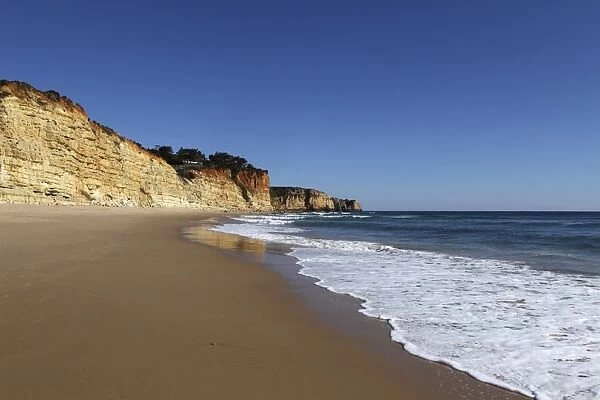 A wave washes an empty beach of golden sand, with steep cliffs, typical of the Atlantic coastline near Lagos, Algarve, Portugal, Europe