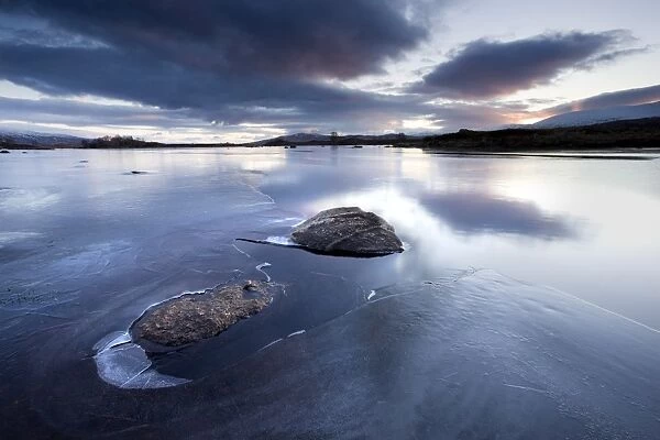Winter view of Loch Ba at dawn, loch partly frozen with two large rocks protruding from the ice, calm morning, Rannoch Moor, near Fort William, Highland, Scotland, United