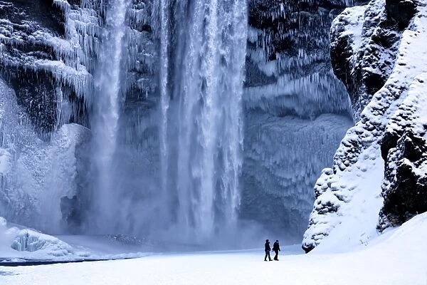 Winter view of Skogafoss waterfall, with cliffs covered in icicles and foregreound covered in snow