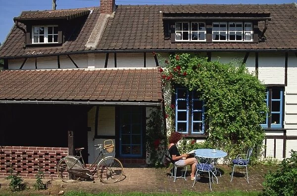 Woman reading at a table outside a typical house, with a bicycle in front
