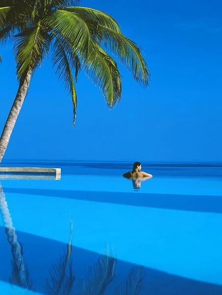 Woman in swimming pool under palm tree looking at sea, Maldives, Indian Ocean, Asia