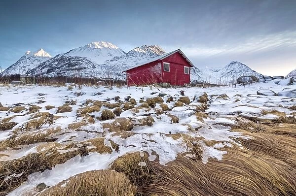 Wood hut framed by rocks covered with grass and ice with snowy peaks in the background