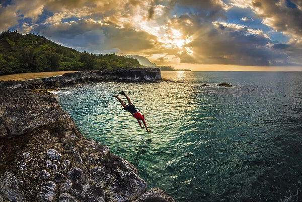 A young man dives into the calm sea on the northern rocky coastline of Kauai at sunset
