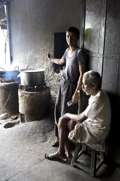 Young man stirring milk for chai, with blind man sitting beside him, roadside chai stall