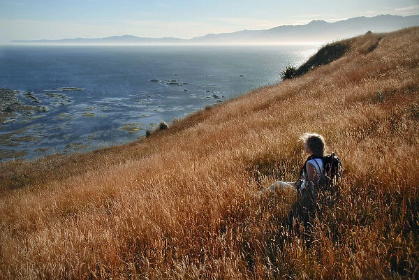 A young woman hiker rests in a wild grass field on the coast of New Zealand